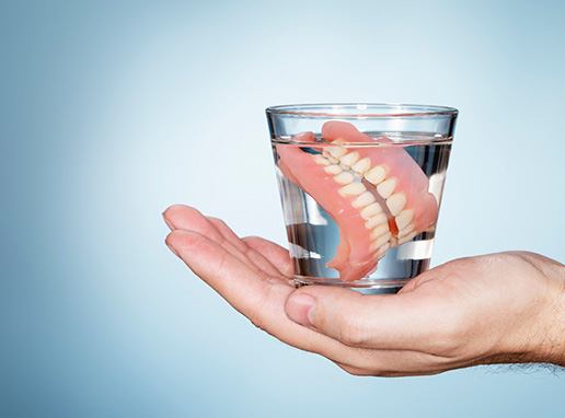 Hand holding a glass with clear fluid and complete set of dentures inside