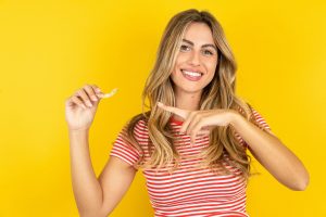 young woman in striped shirt with long blond hair pointing to clear braces in her other hand